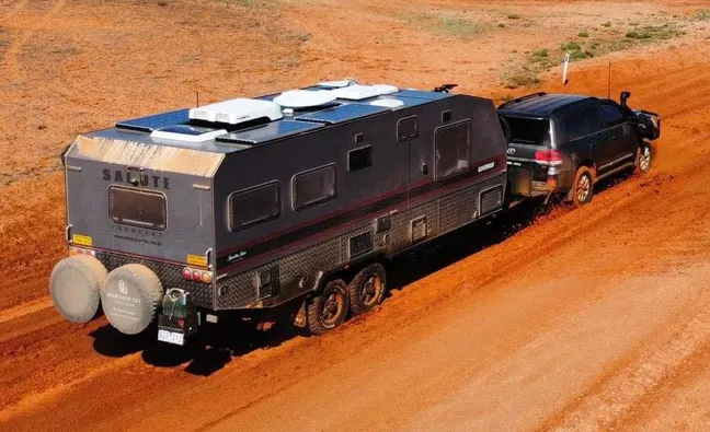 Off-road car towing a large caravan on a dusty dirt track in a remote desert landscape.