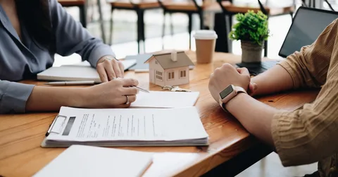 Two people discuss real estate paperwork at a table with a small house model and coffee cup.