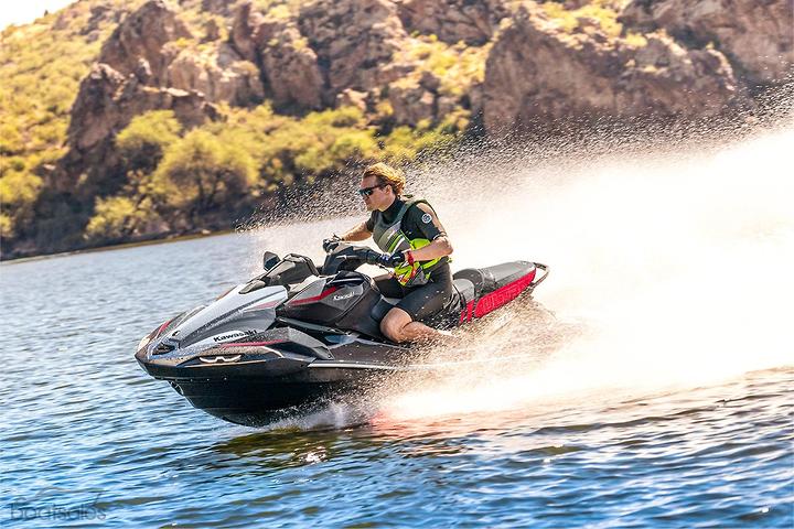 Person riding a jet ski on a sunny day with rocky mountains in the background, creating a splash on the water.
