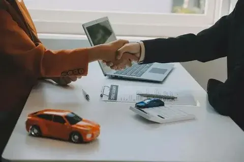 Business negotiation: two people shaking hands over car sale agreement with toy cars and laptop on table.